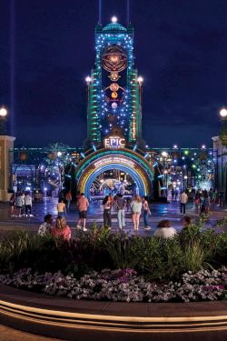 Nighttime scene of a theme park entrance, featuring colorful lights, a tall clock tower, and a crowd of people gathered around a garden.