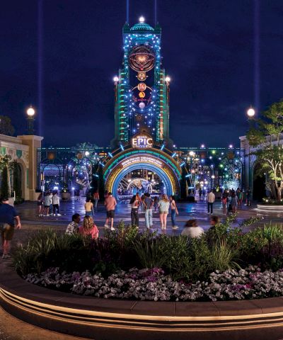 Nighttime scene of a theme park entrance, featuring colorful lights, a tall clock tower, and a crowd of people gathered around a garden.