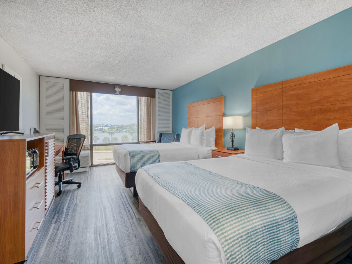 The image shows a hotel room with two beds, a TV, a desk and chair, and a window with a view. The decor is modern and minimalistic.