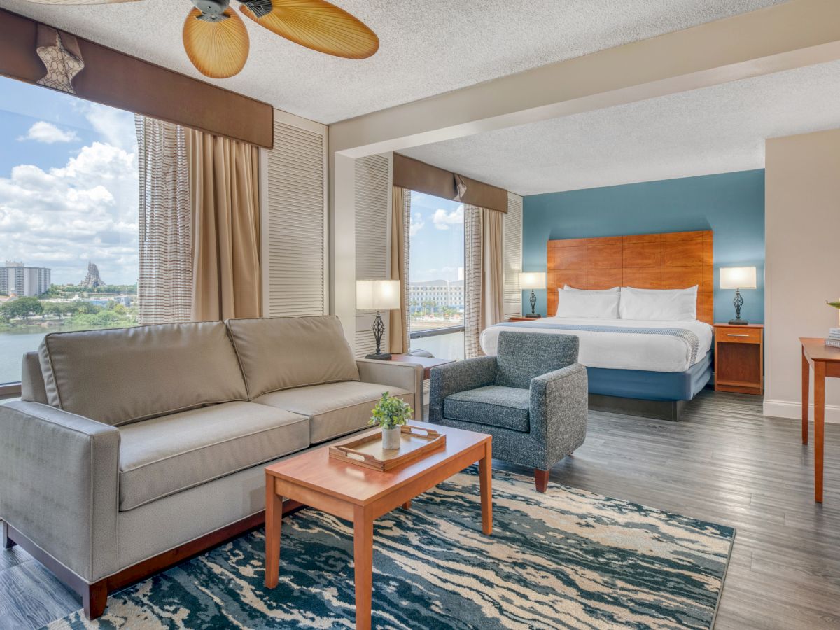 A modern hotel room features a bed, sofa, armchair, table, lamps, ceiling fan, and a window view of a river and city buildings.