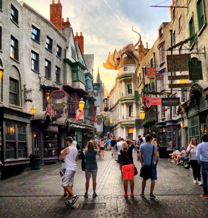 People walking through a themed street with a dragon sculpture above, among various shops and medieval-style buildings, under a partly cloudy sky.