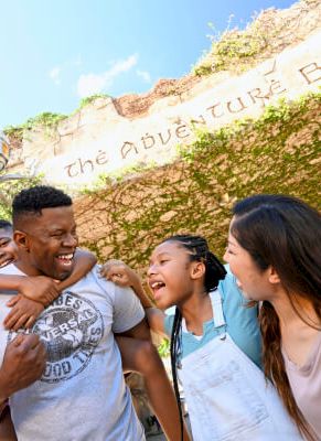 A joyful group stands under an archway with "The Adventure Begins" inscribed, smiling and embracing each other in a lively setting.