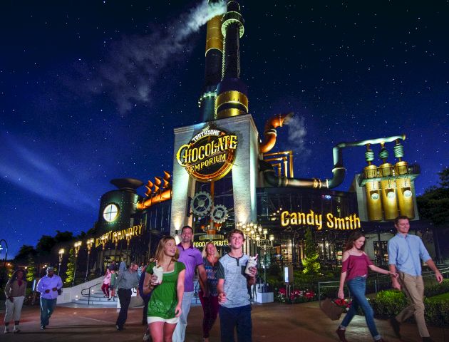 The image shows a whimsical, steampunk-style chocolate factory at night with people walking in the foreground.