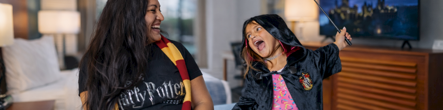A person and a child dressed in Harry Potter attire are joyfully playing on a bed in a hotel room.