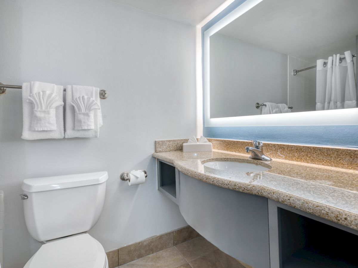 A clean bathroom with a granite countertop, illuminated mirror, sink, toilet, and folded towels on a rack.