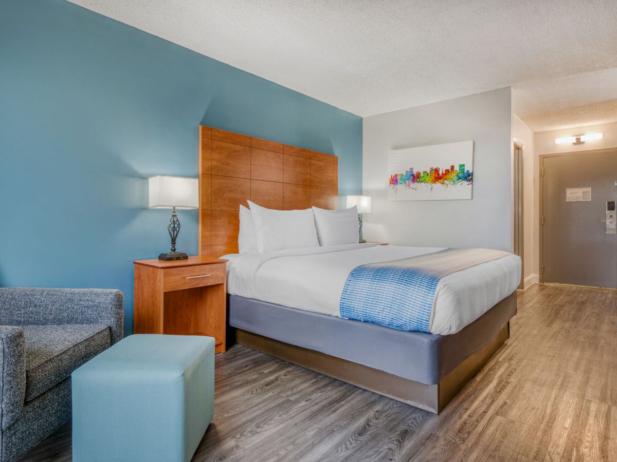 A modern hotel room with a large bed, blue accent wall, artwork, a nightstand, and a small seating area with a chair and ottoman.