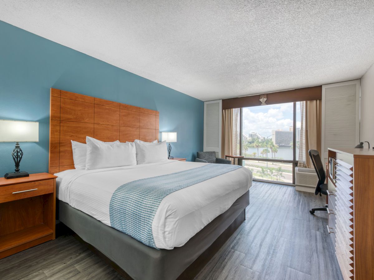 The image shows a modern hotel room with a large bed, side tables, lamps, a desk, and a window offering a view outside.