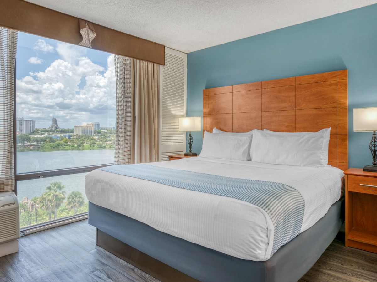 A hotel room with a large bed, bedside lamps, and a window view of a cityscape with a lake and clouds outside.