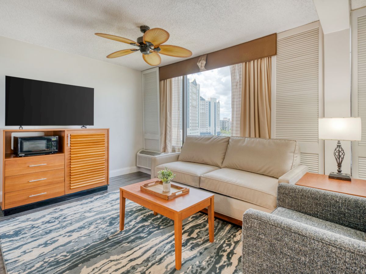 A modern living room with a sofa, chair, TV, microwave, coffee table, lamp, ceiling fan, and window view of a cityscape.