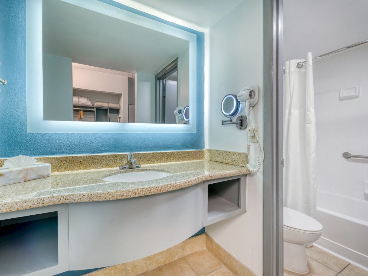 The image shows a bathroom with a lit mirror, sink, countertop, hairdryer, towels, and a shower with a curtain.