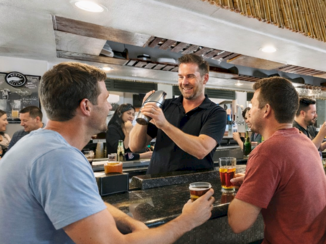 A bartender is mixing a drink while two patrons sit at the bar, each holding a glass, in a lively atmosphere.