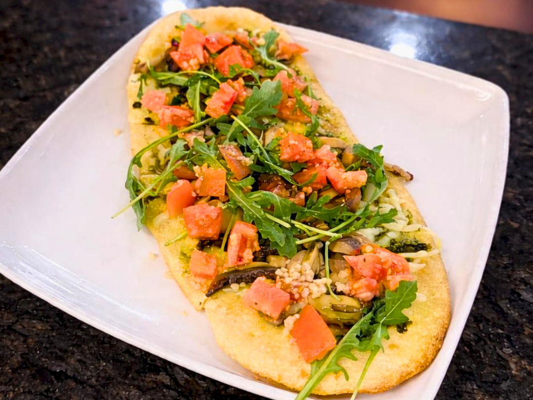 A flatbread topped with arugula, diced tomatoes, and what appears to be cheese sits on a white plate placed on a dark countertop.