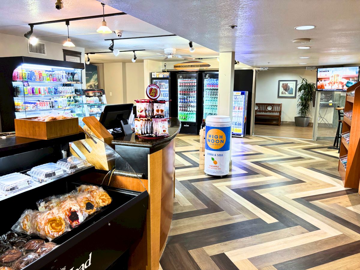 A convenience store with display cases of beverages and snacks, a coffee machine, and assorted products on shelves in a patterned floor setting.