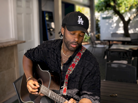 A person wearing a cap plays an acoustic guitar on a patio, surrounded by chairs and greenery, under string lights.