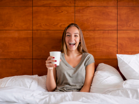 A person sitting in bed with white pillows and holding a coffee cup, smiling in a cozy room with wooden paneling behind.