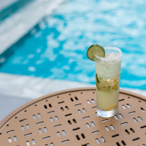 A refreshing drink garnished with a cucumber slice is placed on a round table beside a swimming pool.
