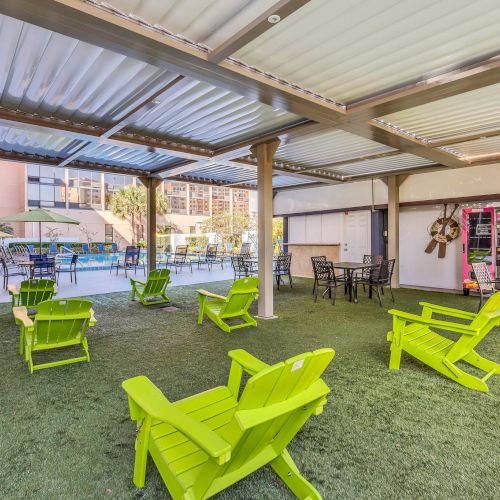 A covered outdoor seating area features bright green chairs on artificial grass, with additional dining tables and chairs, near a swimming pool.