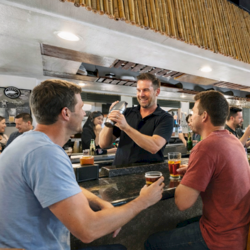 A bartender is shaking a cocktail shaker, serving two men seated at a bar. They hold drinks and are engaged in conversation.