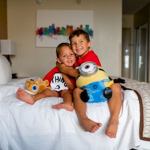 Two children sitting on a bed in a hotel room, smiling and hugging plush toys in matching outfits ending the sentence.