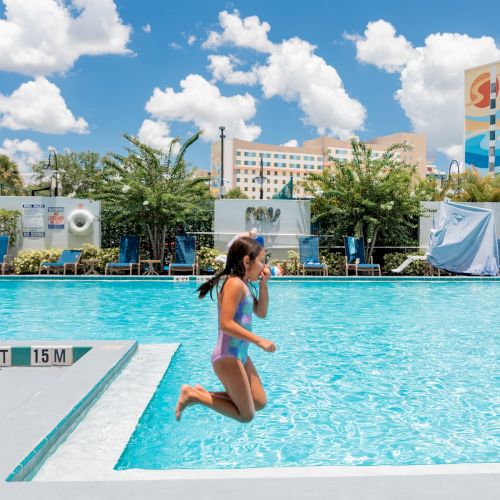 A girl is jumping into a pool at a resort with buildings and greenery in the background, surrounded by poolside chairs and blue skies.