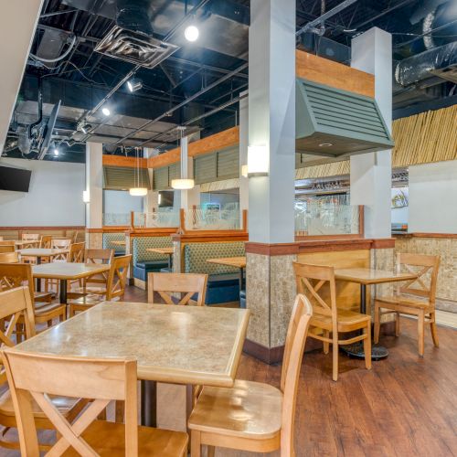 The image shows an empty restaurant interior with wooden tables and chairs, booths, and overhead lighting in a modern setting.
