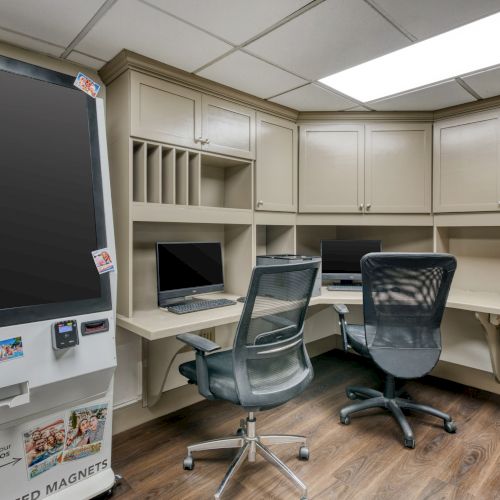 An office space with two computer stations, a large screen or vending machine, and storage cabinets. Chairs and wood flooring are visible.