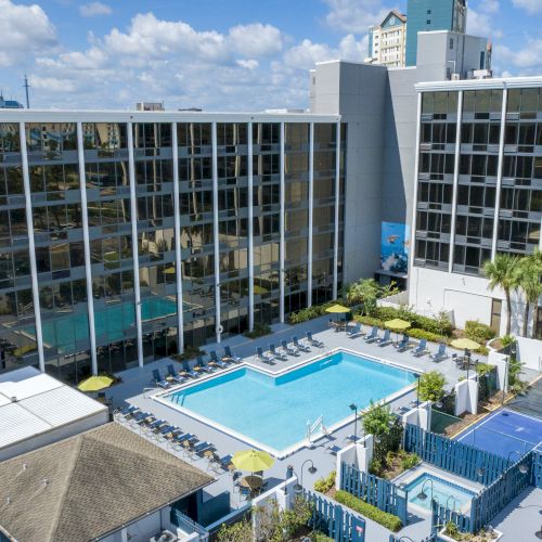 The image shows a hotel with a pool, surrounded by lounge chairs, and a sports court, flanked by buildings and blue skies.