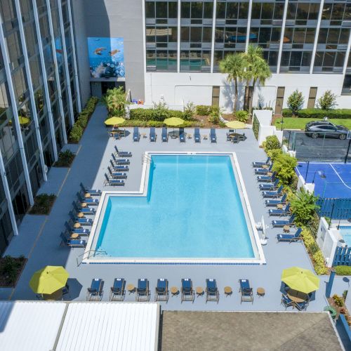 A hotel pool area with loungers, a sports court, and nearby parked cars, surrounded by tall buildings and outdoor greenery.