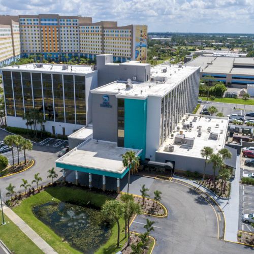 An aerial view shows a large hotel complex with parking lots, roads, and green landscapes under a partly cloudy sky.
