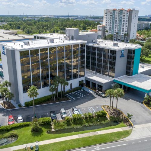 This image shows an aerial view of a multi-story building with a parking lot, surrounded by greenery and roads.