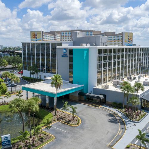 The image shows a hotel with a teal and white facade, surrounded by palm trees and cars in the parking lot, under a partly cloudy sky.