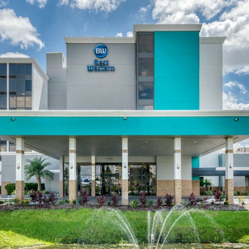 A modern hotel building with a blue and white facade, surrounded by greenery and featuring a small fountain in front.