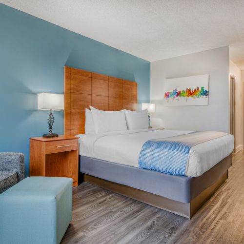 The image shows a hotel room with a large bed, blue walls, a chair, and a lamp on a wooden nightstand.
