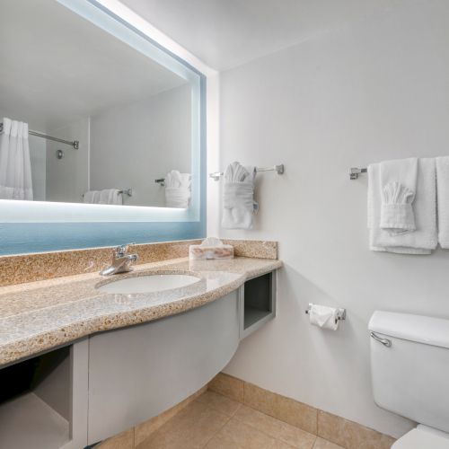 A modern bathroom features a large mirror, a countertop with a sink, neatly folded towels, a toilet, and beige floor tiles.