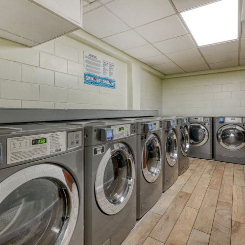 The image shows a laundry room with several industrial washing machines lined up against white tiled walls and a wooden floor.