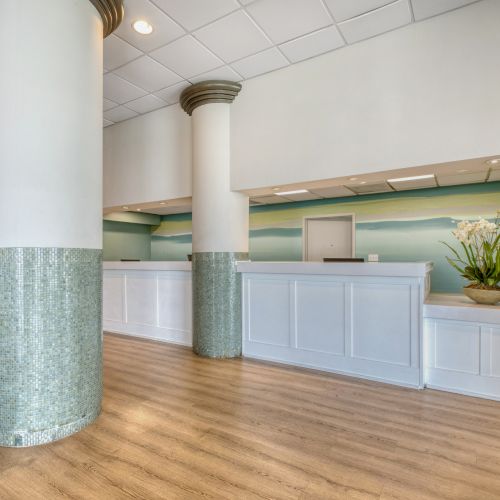 The image shows a modern reception area with white desks, large columns, a wooden floor, and decorative plants on the counter and floor.