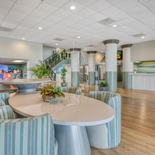 The image shows a lobby area with a long table, striped chairs, plants, and a staircase leading up, with a reception desk in the background.