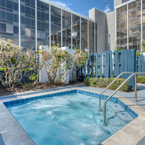The image features a small outdoor jacuzzi with bubbling water, surrounded by plants and a blue fence, beside a tall building.