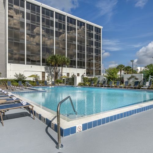 The image shows an outdoor swimming pool surrounded by lounge chairs, with a building and a clear blue sky in the background.