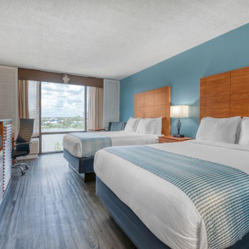 The image shows a hotel room with two beds, a TV, a desk, and a window view. The decor features blue accents and wooden elements.
