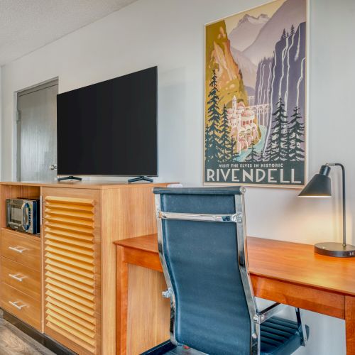A room features a TV, desk, chair, and a Rivendell poster on the wall, with a mirror reflecting part of the space near the doorway.
