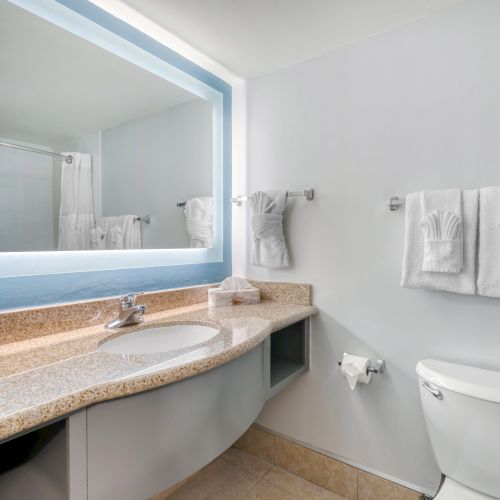 A modern bathroom with a lit mirror, sink, granite countertop, toilet, and neatly folded towels on racks, creating a clean look.