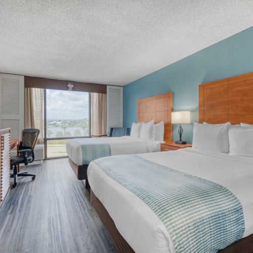 The image shows a hotel room with two beds, a TV, a desk chair, and a window with a view. The decor is modern and clean.