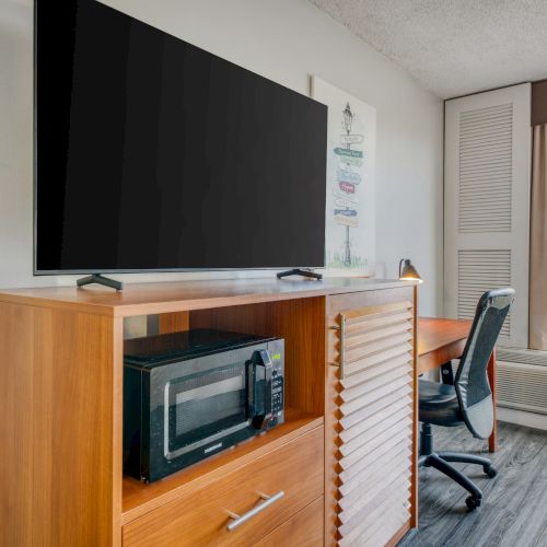 A room features a TV on a wooden stand with a microwave, a desk, a chair, and a window revealing an outdoor scene.