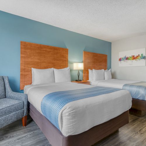 The image shows a hotel room with two beds, a blue accent wall, a chair, a lamp, and colorful artwork on the wall.