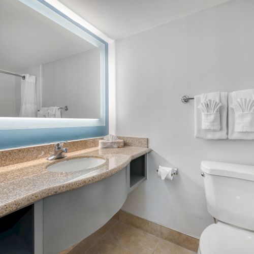 This image shows a modern bathroom with a large mirror, sink, countertop, toilet, and neatly folded towels on a rack.