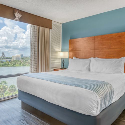 A hotel room with a large bed, two lamps, and a city view through a window overlooking a body of water under a partly cloudy sky.