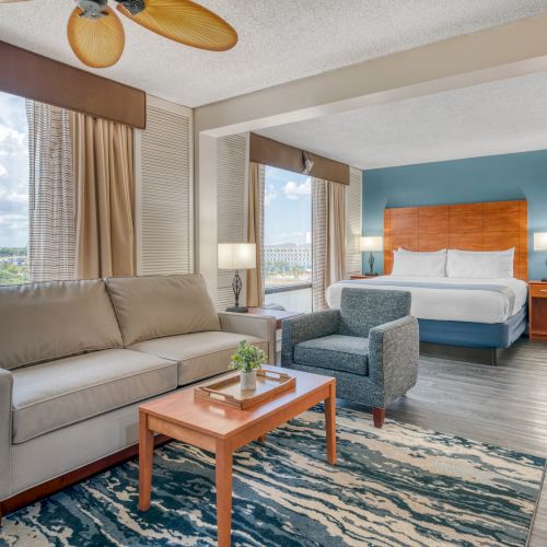 A modern hotel room with a bed, sofa, armchair, and a large window offering a city view.