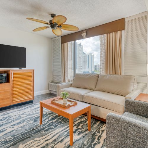 A modern living room with a sofa, armchair, coffee table, TV, ceiling fan, and a large window with a city view.