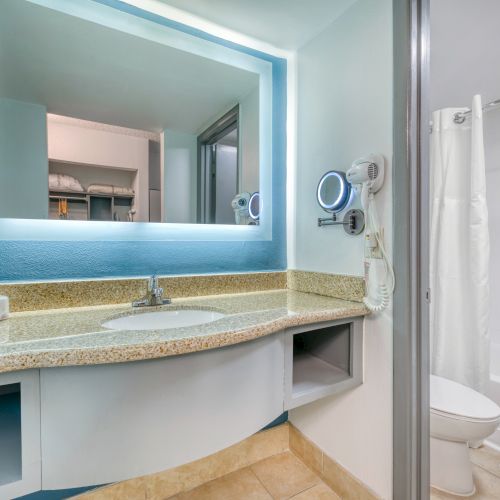 A modern bathroom with a lighted mirror, countertop with a sink, mounted hairdryer, tissue box, and adjacent shower and toilet.
